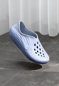 Svetlomodré nazúvacie klobúky s ventilačnými otvormi a logom Nike swoosh, vznášajúce sa nad sivou dlažbou s bielou záclonou v pozadí.