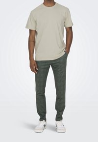T-shirt in cotone beige con scollatura rotonda, abbinata a pantaloni verdi con motivo a quadri. Il modello indossa sneakers bianche con accenti verdi.