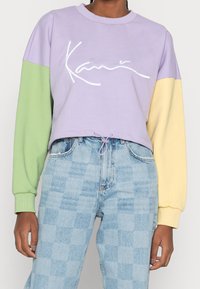 Sudadera corta morada con mangas verdes y amarillas; presenta un logo blanco. Hecha de tela suave con puños acanalados y cordón ajustable.