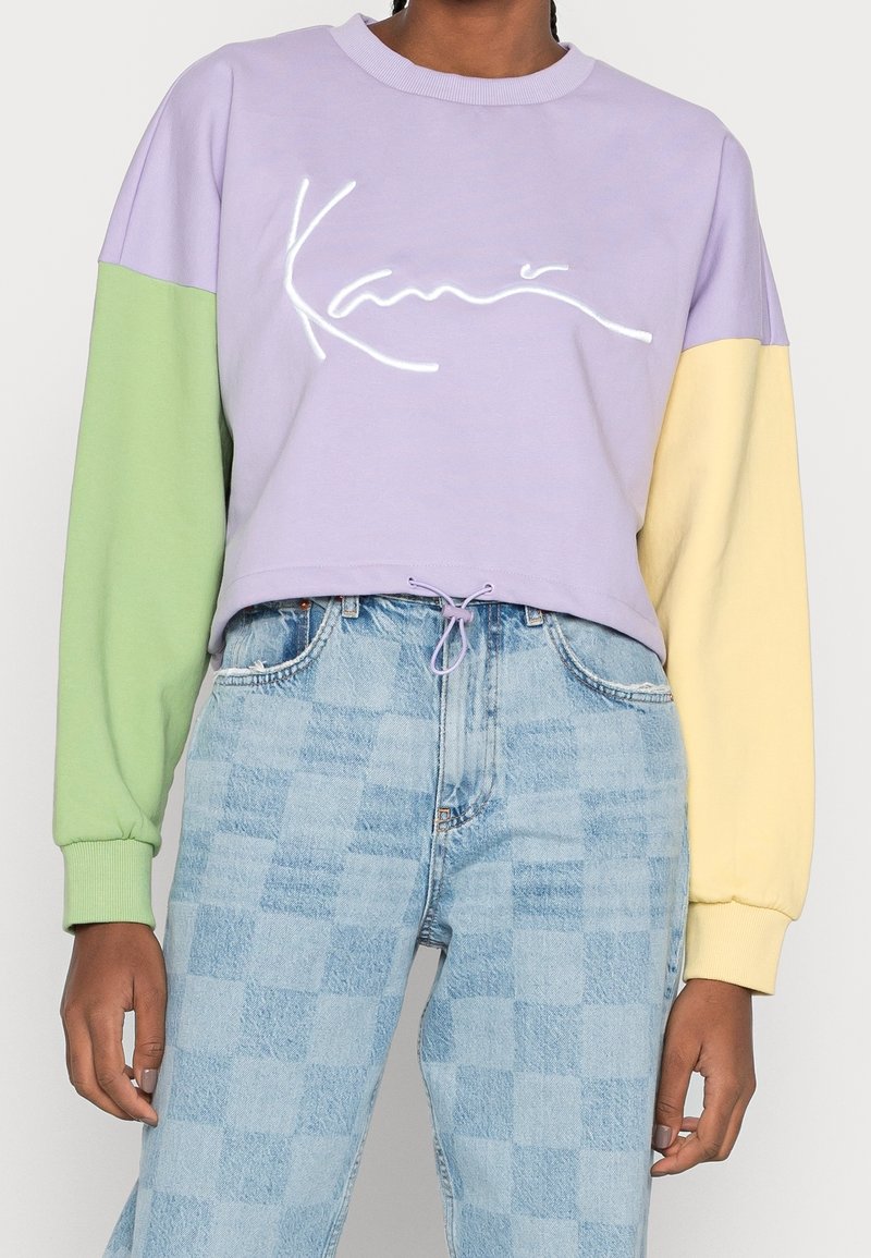 Sudadera corta morada con mangas verdes y amarillas; presenta un logo blanco. Hecha de tela suave con puños acanalados y cordón ajustable.