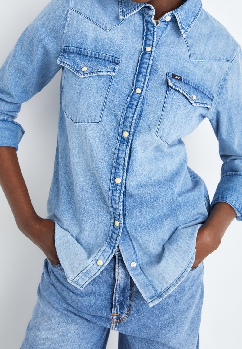 Světle modrá džínsová košile s dlouhými rukávy, dvěma kapsami na hrudi a světlými knoflíky. Tkanina má strukturovaný povrch.