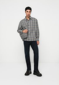 Grijze geruite button-up shirt met lange mouwen, donkere jeans, zwarte chunky schoenen en een grijze schoudertas. Effen witte achtergrond.