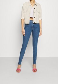 Beige cropped jacket with dark buttons, paired with blue skinny jeans and pink open-toe sandals. Simple design and fitted silhouette.