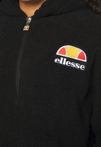 Sudadera de forro polar negra con medio cierre, que presenta un logo bordado multicolor de ellesse en el lado izquierdo del pecho. Textura suave, diseño casual.