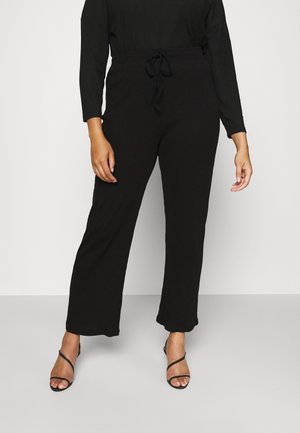 Vrouw draagt zwarte wijde broek met trekkoord in de taille en zwarte sandaal met hak en bandjes, staand tegen een effen lichte achtergrond.