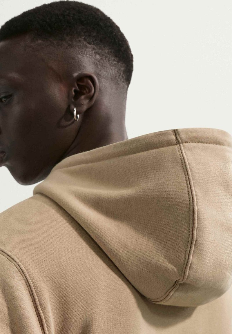Person with short hair and a hoop earring wearing a beige hooded sweatshirt, shown from the side and back against a plain background.