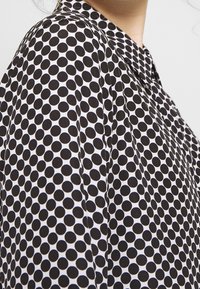 Blouse en tissu blanc à pois noirs, avec un col, portée sur l'épaule et la partie supérieure de la poitrine d'une personne, sur un fond uni.