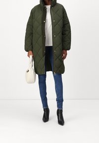 Osoba v dlouhém prošívaném olivově zeleném kabátu, bílém tričku, modrých úzkých džínách a černých kotníkových botách, držící bílou kabelku.