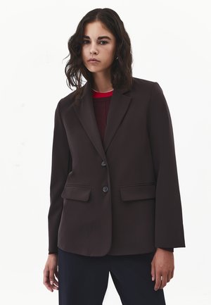 Young woman with shoulder-length dark hair wearing a dark brown blazer over a burgundy top standing against a plain white background.