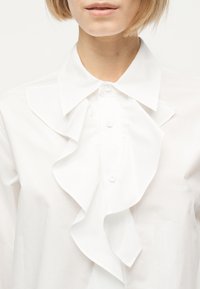 Γυναίκα που φοράει λευκό πουκάμισο με κουμπιά, με φαρδιά γιακά και βολάν στο μπροστινό μέρος, κομμένο στο λαιμό και στους ώμους.