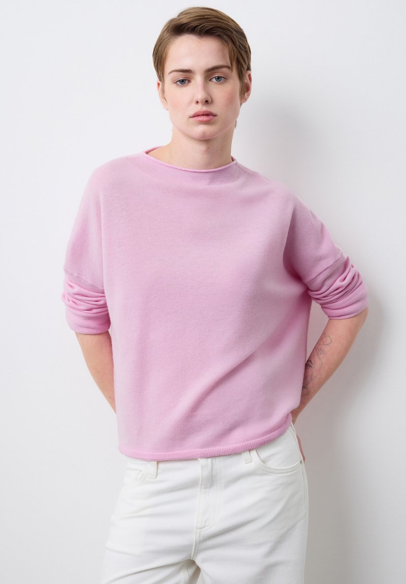 Rosa Pullover aus weichem Stoff mit lockerer Passform, überschnittenen Schultern und aufgerollten Bündchen, kombiniert mit weißen Hosen.