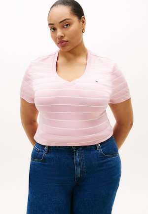 Donna con capelli raccolti che indossa piccoli orecchini a cerchio, una maglietta a righe rosa e bianca con scollo a V e jeans blu, in piedi con le mani dietro la schiena.