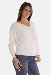 Maglione bianco a scollo a V con maniche lunghe, polsini e orlo a coste. Texture morbida e vestibilità rilassata. Indossato con jeans blu.