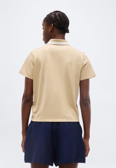 Άτομο με κοτσίδες, γυρισμένο ανάποδα, φοράει μπεζ κοντομάνικη πόλο μπλούζα και σκούρα μπλε σορτς μπροστά σε λευκό φόντο.