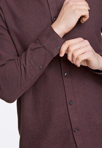 Camicia marrone con bottoni, realizzata in tessuto testurizzato, con colletto, maniche lunghe e bottoni neri con dettagli in filato rosso ai polsini.