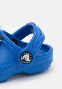 Modré dětské dřeváky s nastavitelným zadním páskem a černobílým knoflíkem s logem krocana.