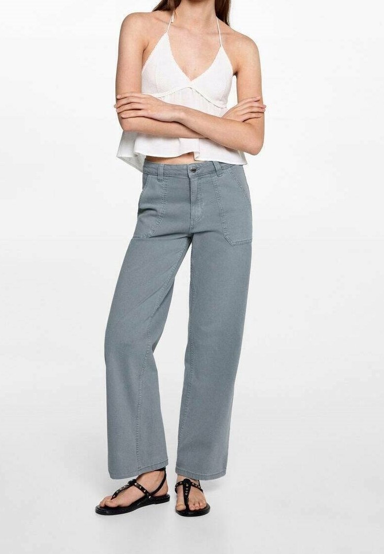 Vrouw met een witte mouwloze haltertop, grijze high-waisted wijde broek en zwarte platte sandalen staat met gekruiste armen.