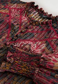 Blouse à motifs rouge, marron et noir avec col à volants, boutons recouverts de tissu, bordure ajourée et poignets élastiqués smockés.
