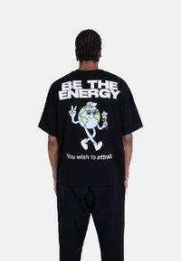 Svart oversized t-skjorte med et livlig grafisk motiv av en tegneserie-jordklode som holder en blomst, med fet tekst over og under designet.