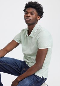Polo shirt verde chiaro con un tessuto testurizzato, dettagli bianchi sul colletto e un logo sul petto, abbinato a jeans scuri.