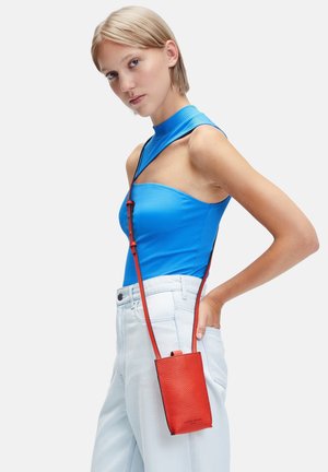 Sac à bandoulière en cuir rouge avec une finition texturée, de forme rectangulaire et une sangle réglable, associé à un haut sans manches côtelé bleu et un jean clair.