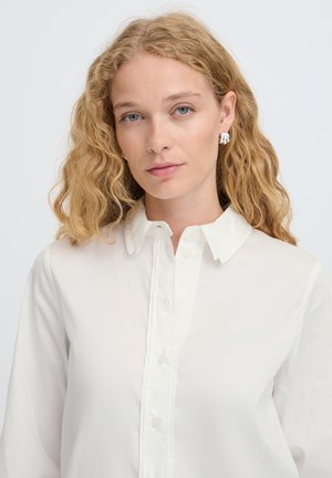 Mujer rubia con cabello ondulado lleva pendientes de aro plateados y una camisa blanca abotonada con cuello sobre un fondo liso y claro.