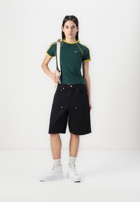 Grønn kortermet topp med gule detaljer, kombinert med svarte shorts til kneet. Merkbare detaljer inkluderer hvite joggesko og lyse sokker.