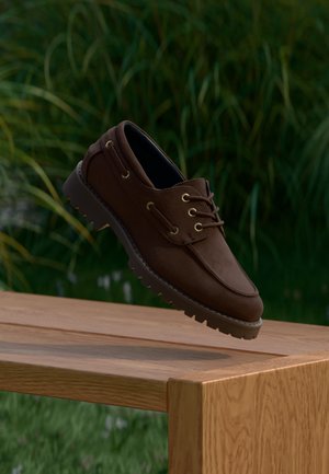 Chaussure bateau en cuir marron à semelle robuste suspendue au-dessus d'un banc en bois, sur fond de feuillage vert extérieur.