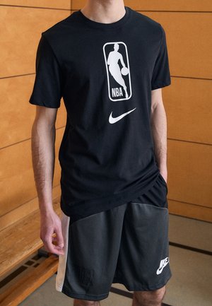 Homme portant un t-shirt noir avec les logos NBA et Nike et un short Nike noir, debout à l’intérieur près d’un banc en bois.