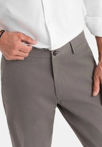 Pantaloni grigi con texture e chiusura a bottone, dotati di frontale piatto e tasche laterali. Abbinati a una camicia bianca con colletto.