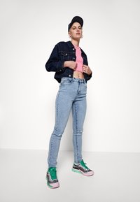 Casaco de ganga, top curto rosa, jeans skinny azul claro e ténis multicoloridos. Apresenta um detalhe desgastado nos jeans e um boné.
