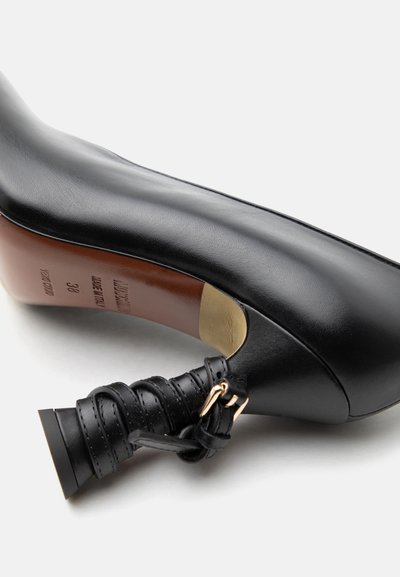 Μαύρο δερμάτινο γυναικείο παπούτσι με ψηλό τακούνι, φαρδύ και τυλιγμένο τακούνι με λεπτομέρεια χρυσής αγκράφας, το οποίο φαίνεται από την πλευρά της σόλας σε λευκό φόντο.