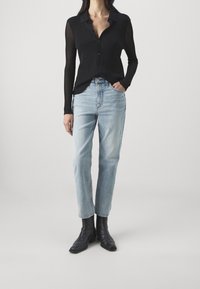 Žena v průhledném černém topu s dlouhým rukávem, světle modrých džínech s vysokým pasem a černých kotníkových botách, stojící s jednou rukou v kapse.