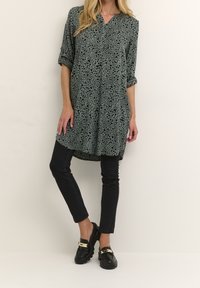 Vestido camiseón en verde con patrón de manchas negras, mangas cortas remangadas, plisado en la parte frontal, dobladillo trasero más largo, combinado con jeans skinny negros y mocasines.