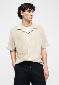 Joven con cabello rizado oscuro que lleva una camisa beige de manga corta abotonada y pantalones negros, mirando hacia un lado con las manos en los bolsillos.