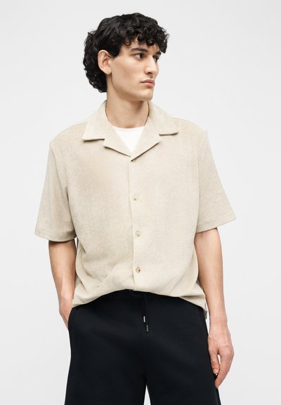 Jeune homme aux cheveux bouclés et foncés portant une chemise beige à manches courtes boutonnée et un pantalon noir, regardant sur le côté avec les mains dans les poches.