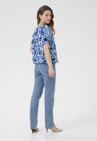 Kurzarmbluse in Blau mit Blumenmuster; kombiniert mit hellblauen Jeans und beigen spitzen Schuhen. Glattes Material, lockere Passform.