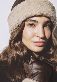 Hnedý strižový klobúk s nadýchaným béžovým pásom, v kombinácii s tmavohnedou koženou rukavicou s textúrovanými akcentmi. Dôraz na teplé farby a mäkkosť.