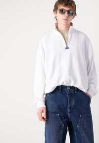 Sweatshirt blanc zippé avec un col haut et des accents bleus, associé à un pantalon cargo en denim bleu avec des poches latérales.