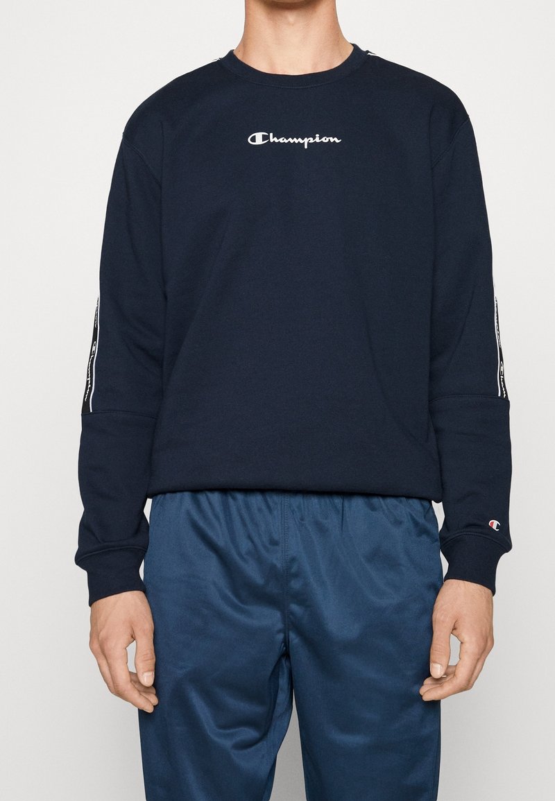 Marine sweatshirt met witte "Champion" logo, ronde hals, lange mouwen en gestreepte accenten op de manchetten, gecombineerd met blauwe sportbroek.