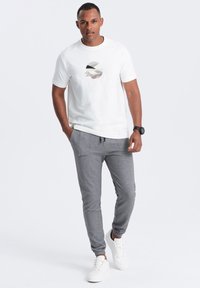 Camiseta blanca con gráfico circular en colores apagados, combinada con pantalones jogger grises con bolsillos laterales y zapatillas blancas. Atuendo casual.