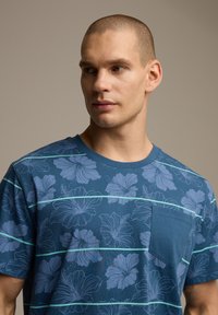 Niebieska koszulka z krótkim rękawem, ozdobiona kwiecistym wzorem w jasnoniebieskim kolorze, z poziomymi paskami i małą kieszonką na lewej piersi.