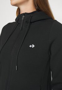 Sweat à capuche noir à fermeture éclair avec un tissu texturé et légèrement extensible. Comprend une capuche, des cordons de serrage et un petit logo argenté sur le côté gauche.