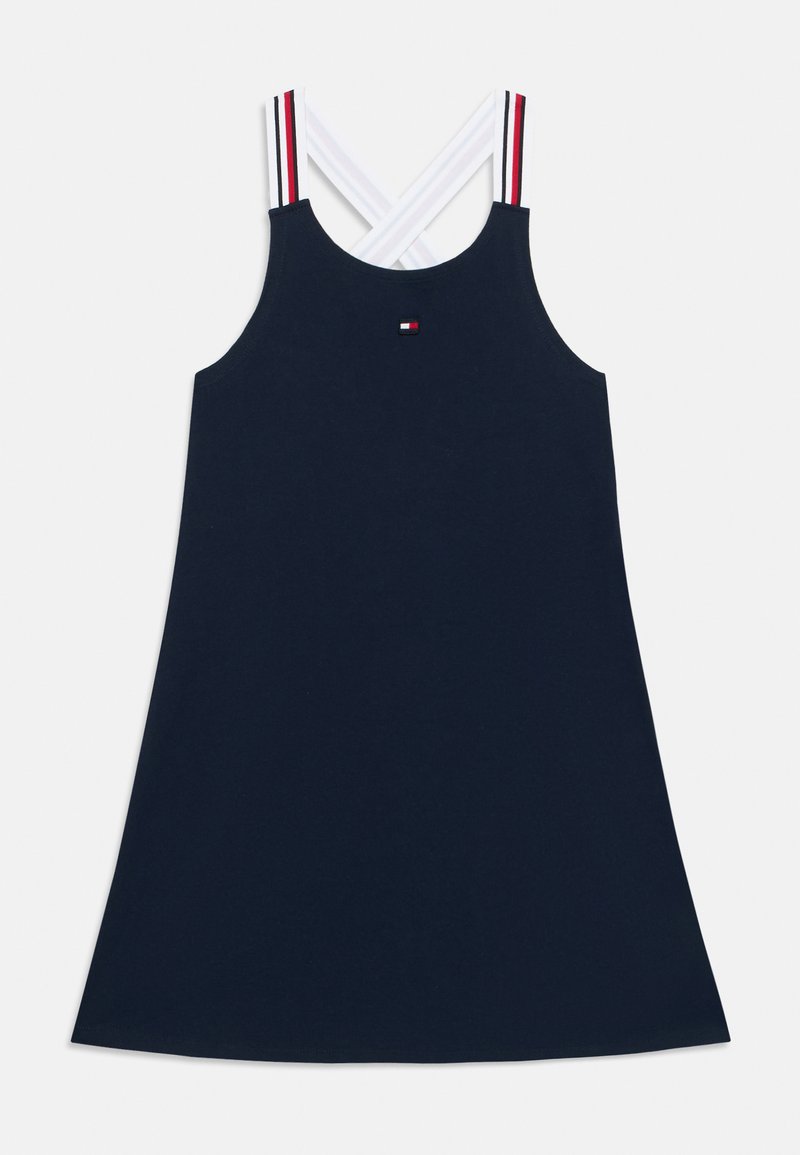 Σκούρο μπλε φόρεμα χωρίς μανίκια με ευθεία σιλουέτα, λευκές διασταυρωμένες τιράντες με κόκκινες και λευκές λεπτομέρειες, και ένα μικρό λογότυπο στο στήθος.