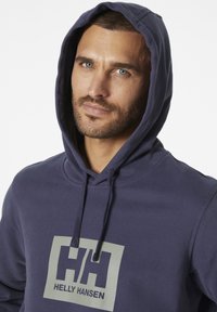 Sudadera de Helly Hansen en azul marino con un bolsillo canguro en la parte delantera, capucha ajustable con cordón y gran logo gris en el pecho. Textura de tela suave.
