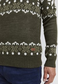 Pull en maille vert foncé avec des motifs floraux blancs et olive, soufflets et ourlet côtelés, avec une petite étiquette marron à l'avant.