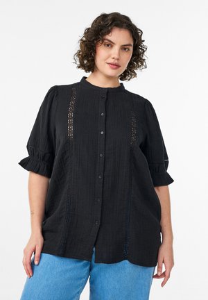 Kobieta z kręconymi włosami nosi czarną bluzkę na guziki z koronkowymi detalami i bufkami na rękawach, połączoną z jasnoniebieskimi dżinsami, na białym tle.