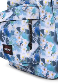 Batoh Eastpak s květinovým vzorem v bílé a žluté barvě na modrém pozadí. Obsahuje černou nášivku s logem a stahovací zip.