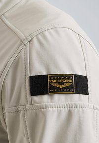 Beige jacket shoulder featuring quilted texture, visible stitching, and a black fabric label with gold and white text detailing the brand.