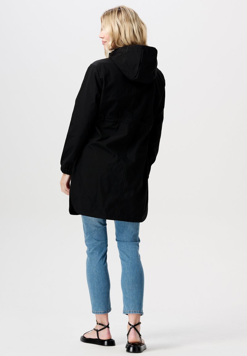 Schwarze wasserdichte Jacke mit Kapuze, tailliertem Schnitt und langen Ärmeln, kombiniert mit hellblauen Skinny-Jeans und flachen schwarzen Sandalen.
